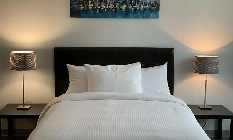 A neatly made bed with white linens and pillows stands between two bedside tables, each with a lamp. Above the bed hangs a colourful cityscape painting on the wall.