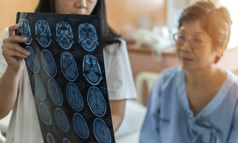 Une professionnelle de la santé tient et examine des images radiologiques du cerveau dans un cabinet. Une personne âgée en jaquette d’hôpital est assise à côté d’elle et l’observe attentivement.
