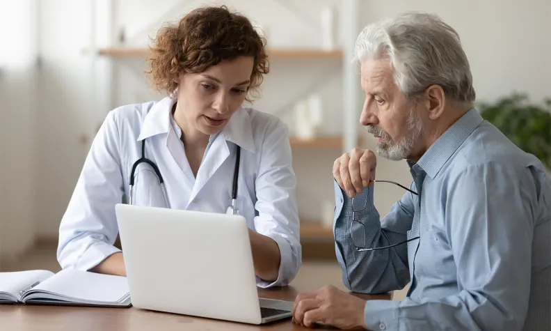 Une professionnelle de la santé en blouse blanche montre des informations sur un ordinateur portable à un homme âgé aux cheveux gris avec une barbe grise. Tous deux sont assis à un bureau et semblent concentrés sur leur discussion.