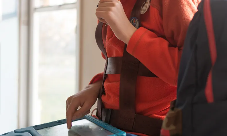 Une personne vêtue d’une veste polaire orange utilise une tablette avec une main tout en mâchouillant un crayon. La personne porte un harnais et se tient près d’une fenêtre où la lumière du jour entre à flots.
