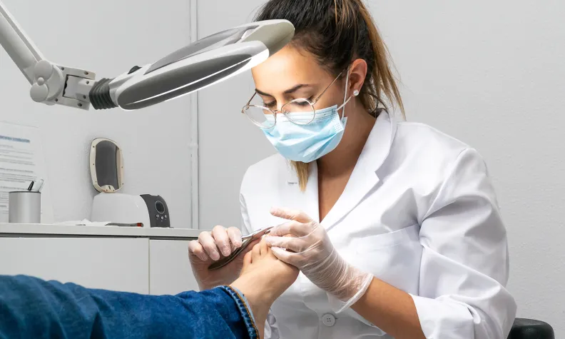 Une podologue portant un masque, des gants et une blouse blanche soigne le pied d’une personne sous une lampe d’examen dans un cabinet.