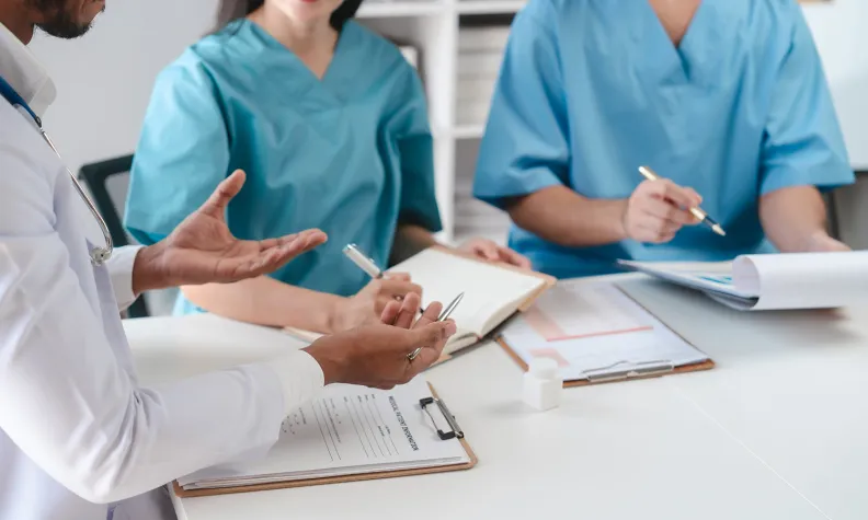 Trois professionnels de la santé en uniforme médical sont assis autour d’une table. Ils discutent de notes et tiennent des stylos. Il y a des documents médicaux et des blocs-notes devant eux.