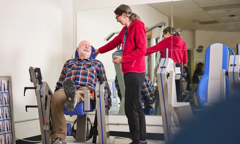 Une personne âgée utilise un appareil d’entraînement pour les jambes tandis qu’une personne en chandail à capuchon rouge lui sourit dans une salle de sport ou une salle de physiothérapie équipée de miroirs et d’appareils d’exercice.