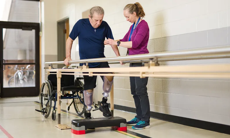 Une physiothérapeute aide une personne âgée munie de prothèses de jambes à s’entraîner à marcher avec des barres parallèles dans un centre de réadaptation. Il y a un fauteuil roulant à proximité.