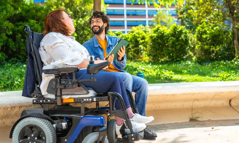 A person in a motorized wheelchair and a person holding a tablet sit on a curved bench outdoors, smiling and talking with greenery and a building in the background.