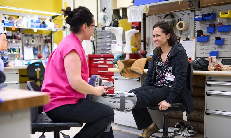 Deux personnes sont assises face à face dans un atelier. L’une porte une botte orthopédique et sourit. L’autre, en chandail rose, examine ou ajuste la prothèse. En arrière-plan, on aperçoit des outils et des pièces de prothèse.