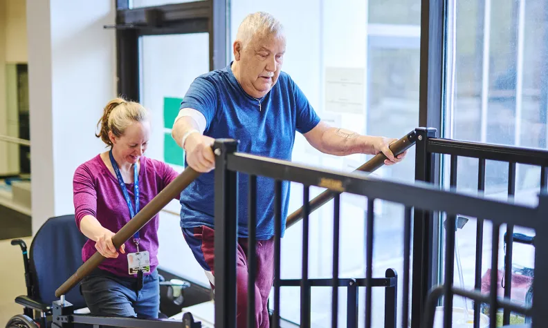 Un physiothérapeute aide une personne âgée pendant qu’elle s’exerce à monter une petite série de marches thérapeutiques, en utilisant les rampes pour se soutenir dans un centre de réadaptation.