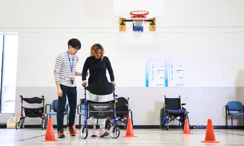 Une personne marche à l’aide d’une marchette en suivant un parcours de cônes orange dans un gymnase. La personne est soutenue par une autre personne à ses côtés. Des fauteuils roulants sont alignés le long du mur sous un panier de basketball.