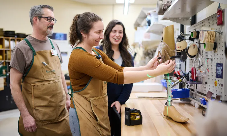 Trois personnes en tablier se tiennent devant un établi dans un atelier, souriantes, et travaillent à l’assemblage d’une prothèse de jambe. Des outils et du matériel sont rangés sur le mur et dans l’étagère en arrière-plan.