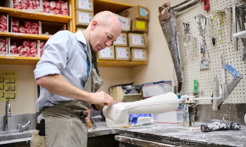 Une personne en tablier façonne un moulage de jambe en plâtre dans un atelier. La personne est entourée d’outils et d’étagères chargées de boîtes. Il y a de la poussière de plâtre sur le comptoir.