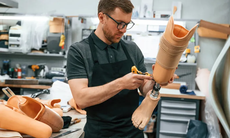 Un homme portant des lunettes et un tablier travaille à l’assemblage d’une prothèse de jambe dans un atelier rempli d’outils et de prothèses