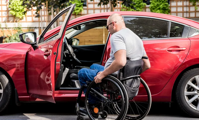 Une personne en fauteuil roulant se prépare à monter dans le siège conducteur d’une voiture rouge dans un stationnement pour personnes à mobilité réduite. La porte du conducteur est ouverte, et il y a la verdure en arrière-plan.