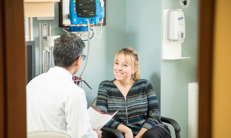 A healthcare professional holding a piece of paper, is seated across from a seated person in a medical setting and engaging in a conversation.