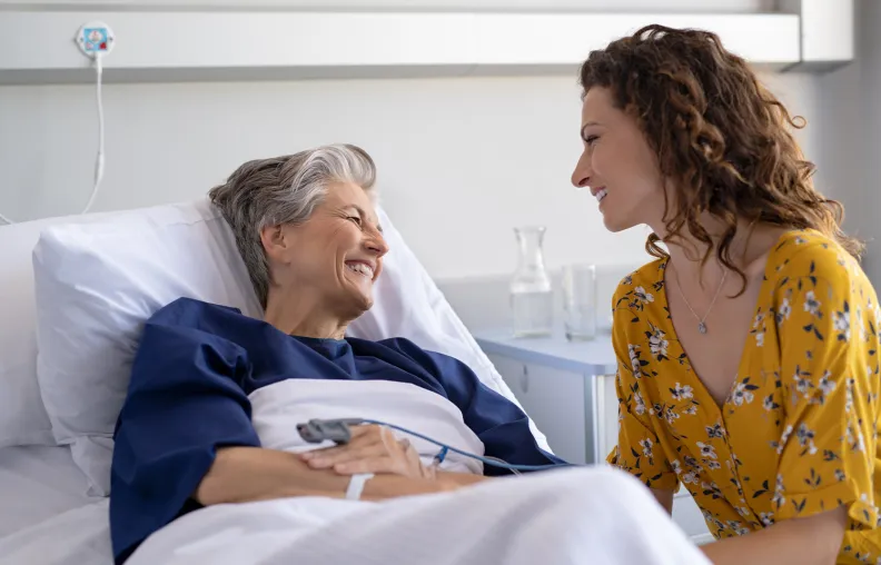 A smiling elderly person lies in a hospital bed, holding hands and sharing a happy moment with a younger person in a yellow floral blouse sitting beside them.
