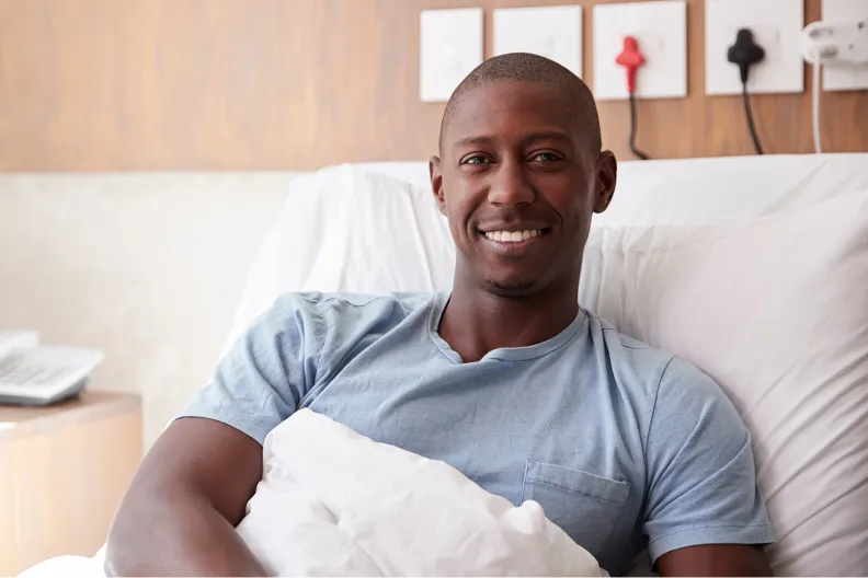 A patient wearing a light blue T-shirt smiles while sitting up in a hospital bed, covered with a white blanket. The background shows a wooden wall with electrical outlets and a telephone on a side table.