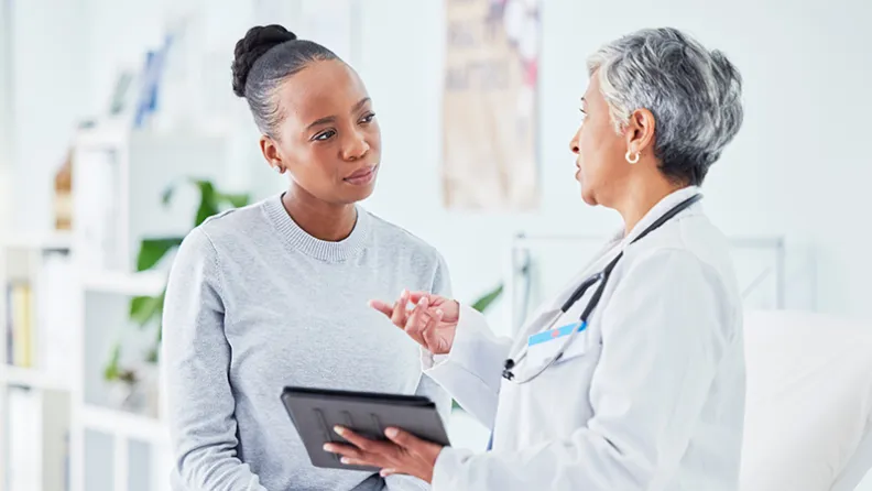 Une médecin en blouse blanche tient une tablette et parle avec une patiente assise sur une table d’examen dans un bureau de consultation lumineux.