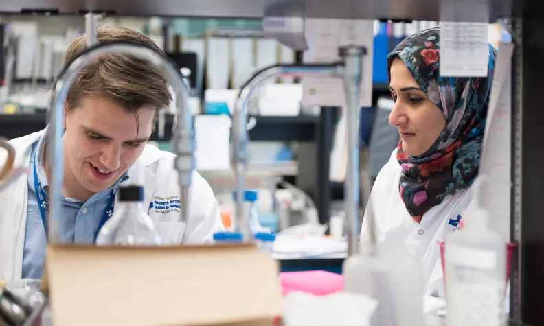 Two individuals wearing white lab coats work together in a busy laboratory setting. 