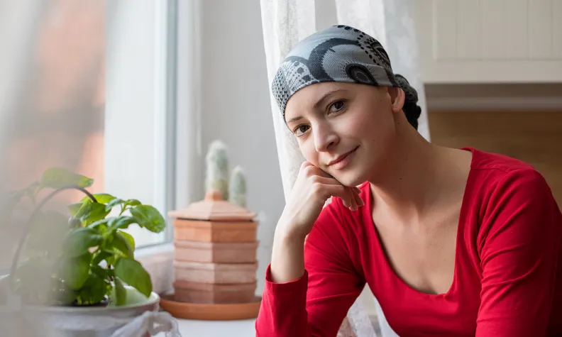 Une personne portant un foulard à motifs est assise près d’une fenêtre lumineuse, la tête reposant sur une main.