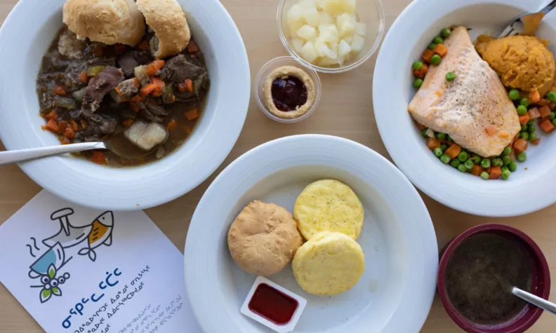 Overhead view of a hospital meal featuring traditional foods, including stew with bannock, salmon with vegetables, soup, and side dishes arranged on a table.