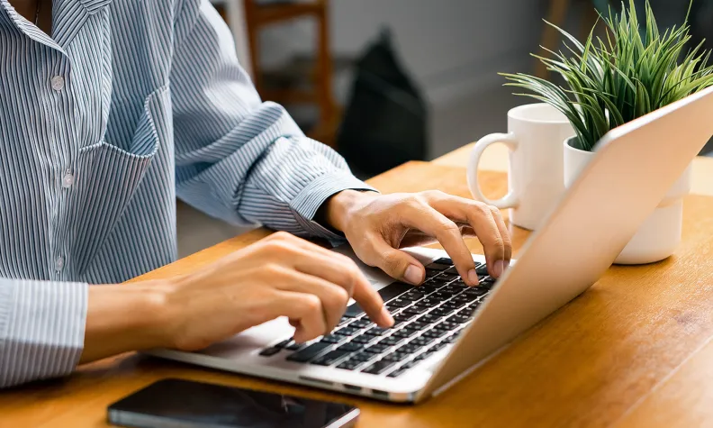 A person typing on a laptop at a wooden desk.