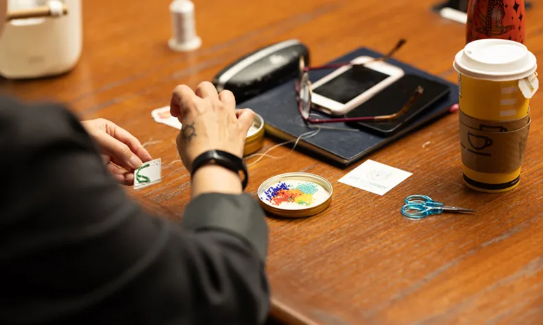 A workshop participant sewing green beads onto her pattern. The workspace in front of her is cluttered with beads, scissors, white thread, a notepad, glasses, phones and a disposable cup of tea.