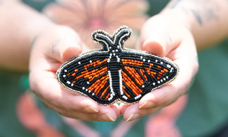 Two hands holding a beadwork monarch butterfly, made of black, white and orange beads. 