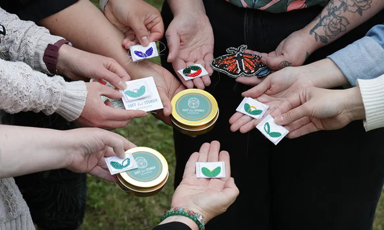 A group of people standing in a circle, holding out their beaded creations, as well as the take-home kits prepared by workshop facilitator, Kaytlyn.