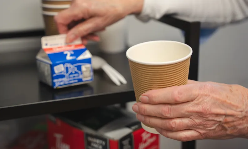 Gros plan sur la main gauche d’une femme âgée tenant une tasse de café. En arrière-plan, sa main droite tient un pot contenant du lait.