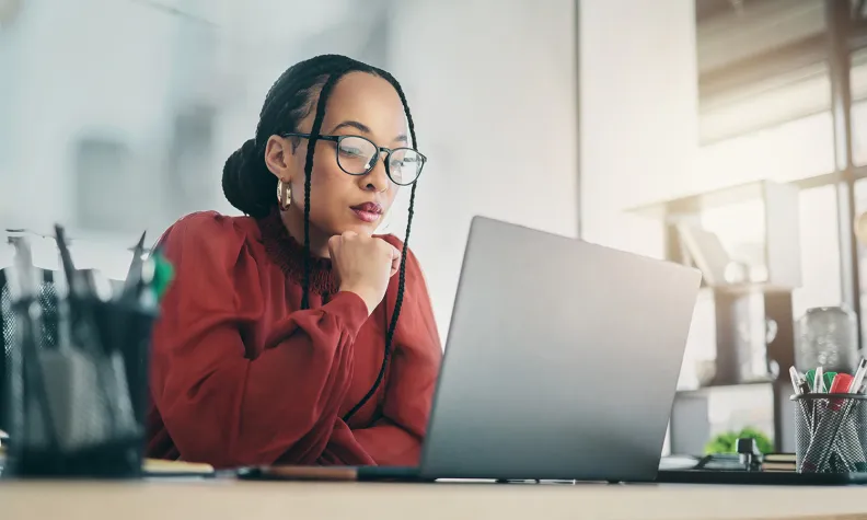 Person wearing glasses sits at a desk using a laptop, resting their chin on one hand, with office supplies and a softly lit workspace in the background.
